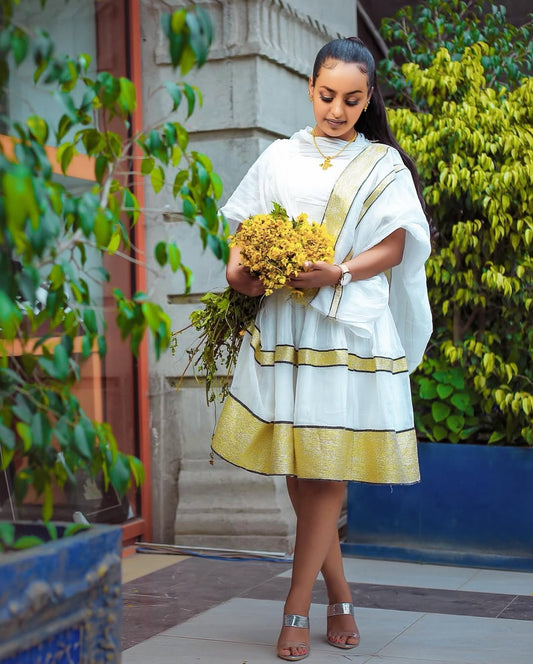 Woman in a white and gold traditional outfit holding yellow flowers Simple Short Golden Habesha Dress Modern Summer Ethiopian Attire ethgebya gebeya usa habesha kemisoutdoors.