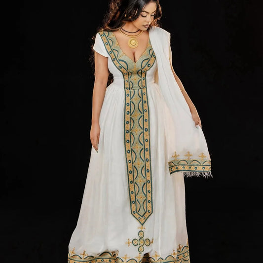 Woman wearing a long, white dress with green and gold embroidery on a black background. Green & Gold Zuria Habesha Kemis Ethgebya
