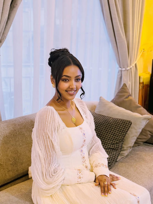Woman in a white outfit sitting on a couch with curtains in the background Long Sleeve Habesha Kemis | New Minimalist Ethiopian Traditional Dress ethgebya