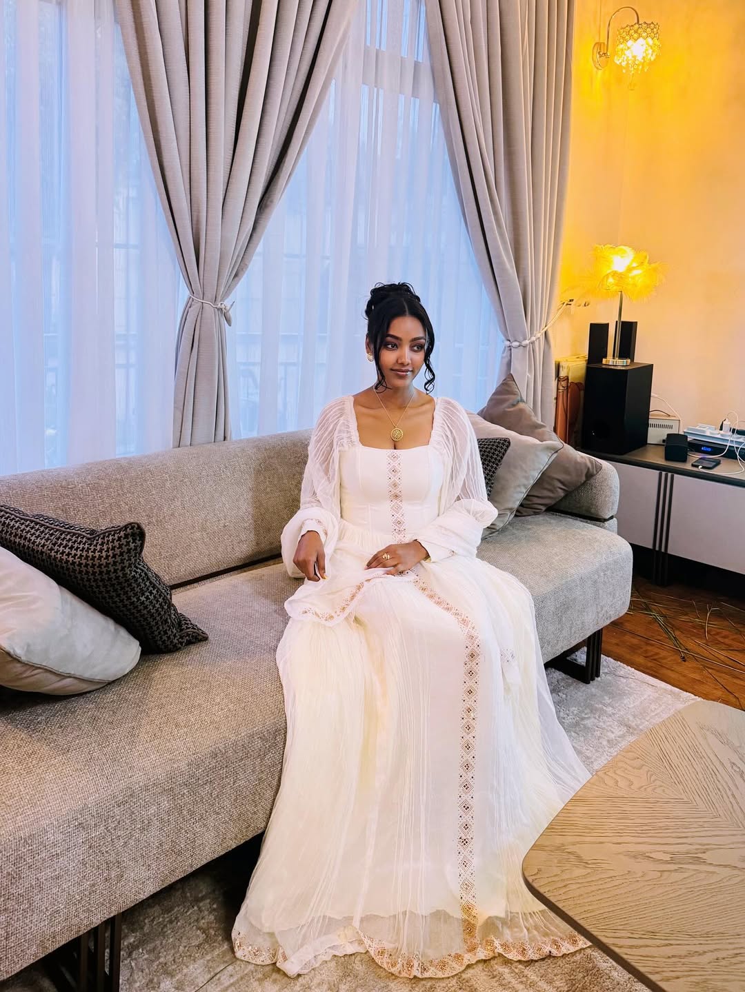 Woman in a white dress sitting on a gray sofa in a well-lit room. Long Sleeve Habesha Kemis | New Minimalist Ethiopian Traditional Dress ethgebya