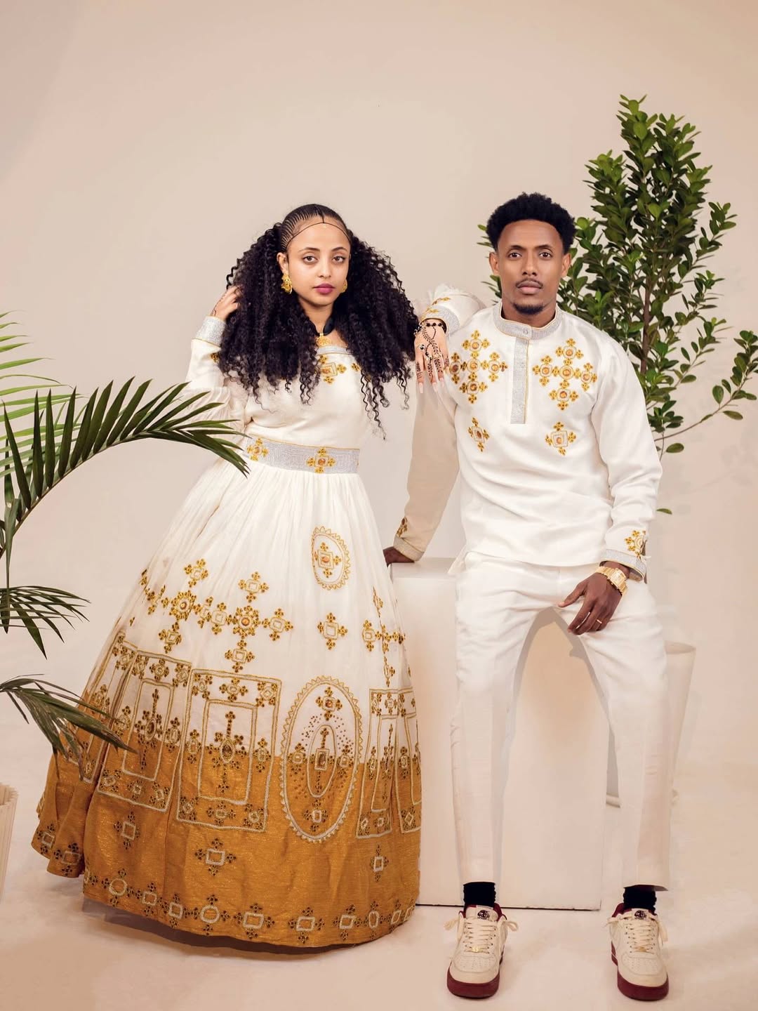 Couple in traditional attire with gold embroidery, standing against a neutral background with plants. Ethiopian gown, Habesha wedding dress, Eritrean evening dress, Ethiopian traditional gown, Ethiopian heritage dress, Habesha chic, Eritrean cultural attire, Habesha elegant dress, Eritrean bridal wear, Ethiopian vintage dress, Habesha bridesmaid dress, African ethnic dress, Ethiopian handcrafted dress, Eritrean handcrafted dress, traditional African gown, Habesha festive dress, Ethiopian formal wear,