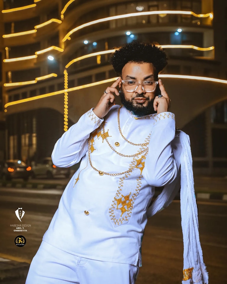 Man in a white embroidered outfit with gold details standing in front of a illuminated building at night. Habesha inspired fashion, Ethiopian custom made dress, Habesha modern Kemis, bridal dress, wedding outfit, Ethiopian embroidered gown, ceremony dress, Habesha festive dress, Ethiopian cultural gown, elegant attire, Habesha handmade , heritage dress, festive gown, Eritrean festive outfit, lace Kemis, designer dress, artisan clothing, festive gown, premium , traditional cotton attire, embroidered gown, ha
