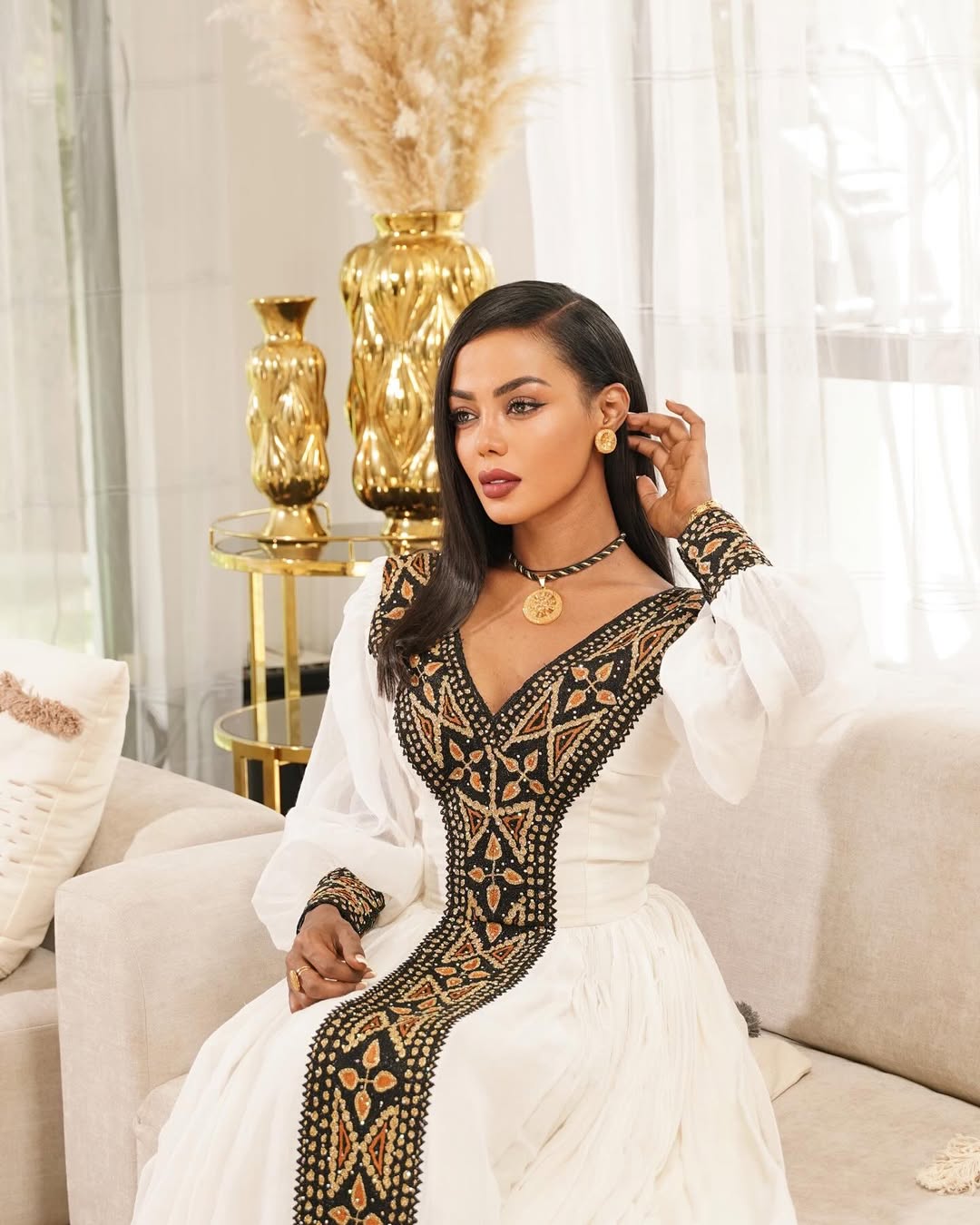 Woman in a white dress with black embroidery sitting in a stylish room. Black & Gold Tilet Zuria Habesha Kemis New Modern Ethiopian Dress ethgebya gebeya habesha kemis zuria kemis
