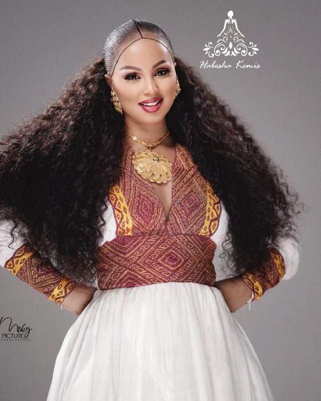 Woman with long, flowing hair wearing a traditional outfit with intricate patterns on a gray background. Modern Zuria Habesha Kemis Handwoven Ethiopian & Eritrean Dress Ethiopian Gebeya Ethgebya