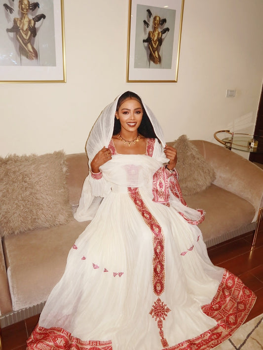 Woman in a white and red embroidered dress sitting on a couch with framed artwork in the background. New Pink Zuria Habesha Kemis Handcrafted Ethiopian Traditional Dress Ethiopia Gebebya Ethgebya