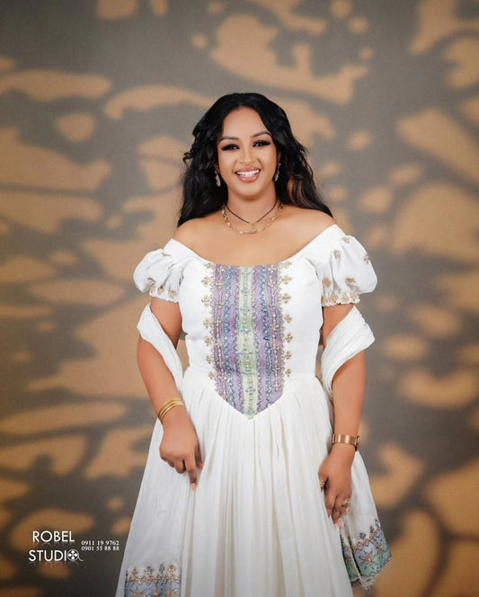 Woman in a white dress with colorful patterns against a patterned background. Modern Luxury Zuria Kemis Elegant Ethiopian Traditional Dress ethgebya gebeya
