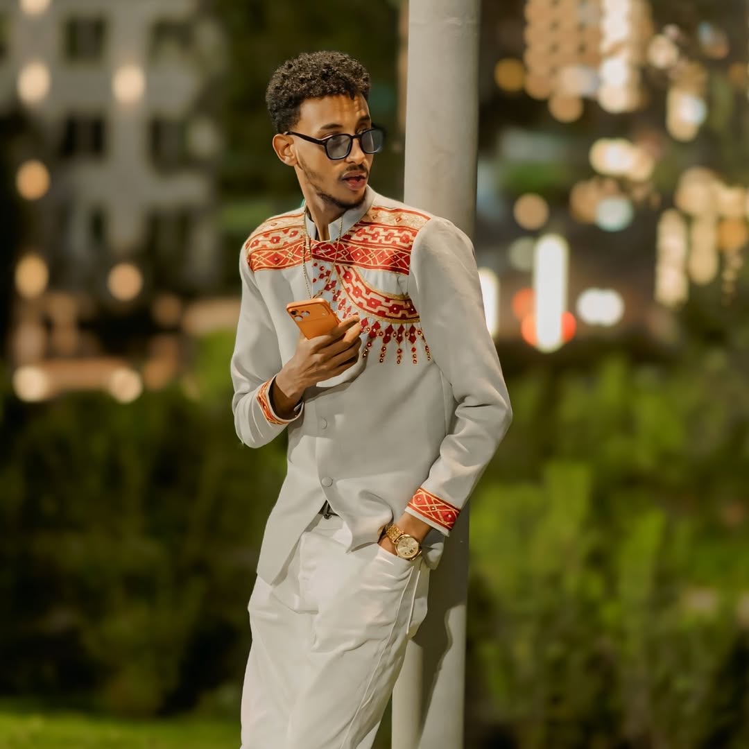 Man in a white outfit with red patterns using a phone outdoors at night. Handwoven Red Habesha Men’s Shirt Ethiopian Traditional Outfit ethgebya ethiopian gebeya