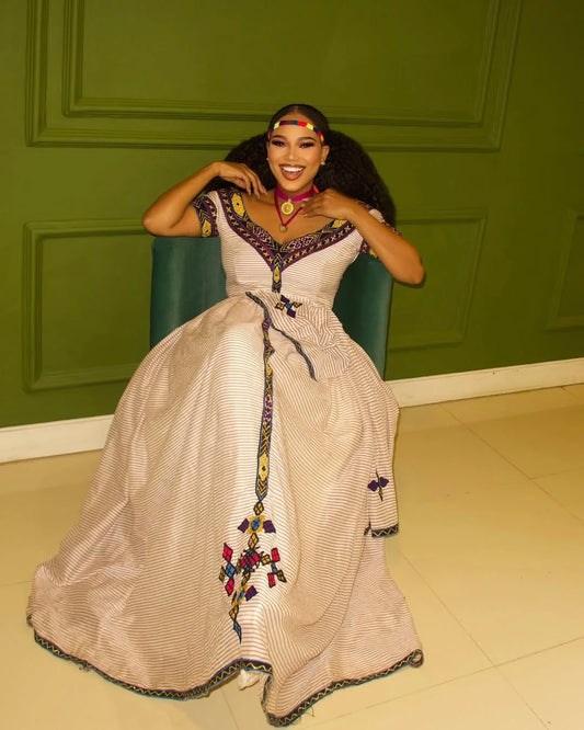 Woman in a decorative dress sitting against a green wall. New Striped Zuria Kemis | Modern Handcrafted Ethiopian Dress