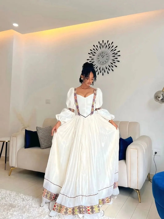 Woman in a white dress with colorful details standing in a modern living room. One of a Kind Modern Zuria Kemis | Handcrafted Ethiopian Dress
