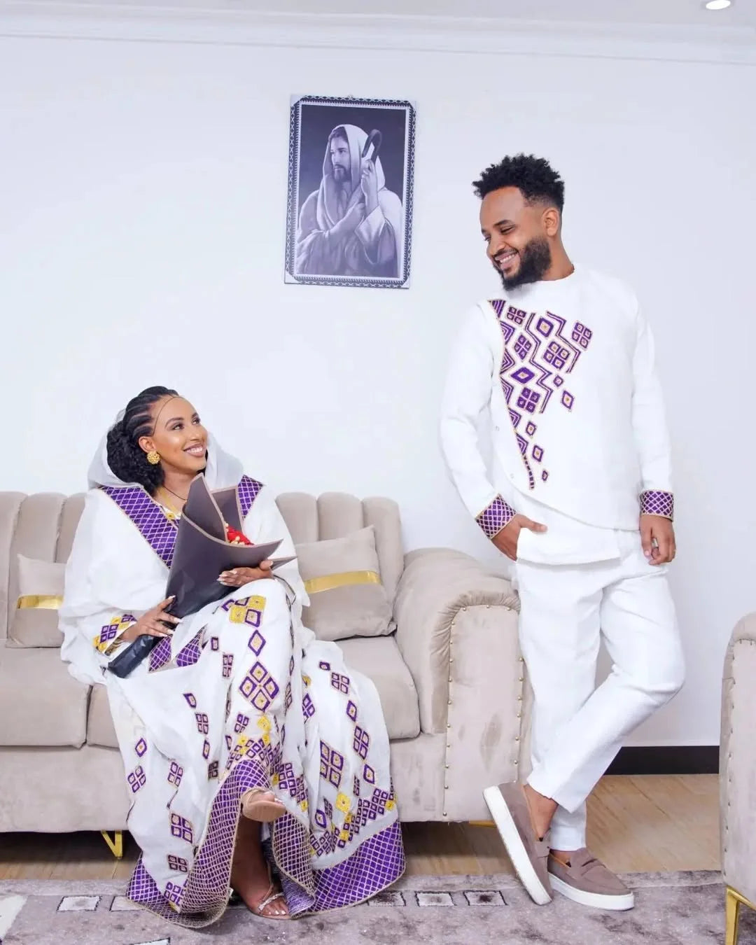 Two people in matching white and purple outfits sitting on a couch in a room with a framed picture on the wall.  Purple Habesha Couple Outfit New Matching Ethiopian Traditional Set ethgebya gebeya
