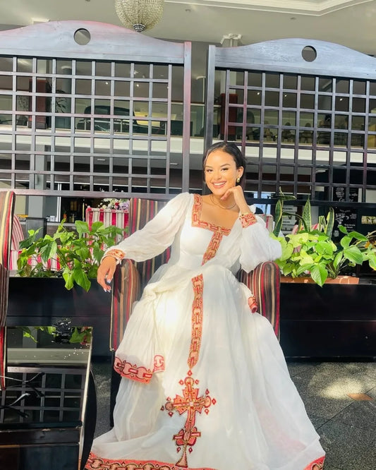 Woman in a white dress with red embroidery sitting in a decorated room.