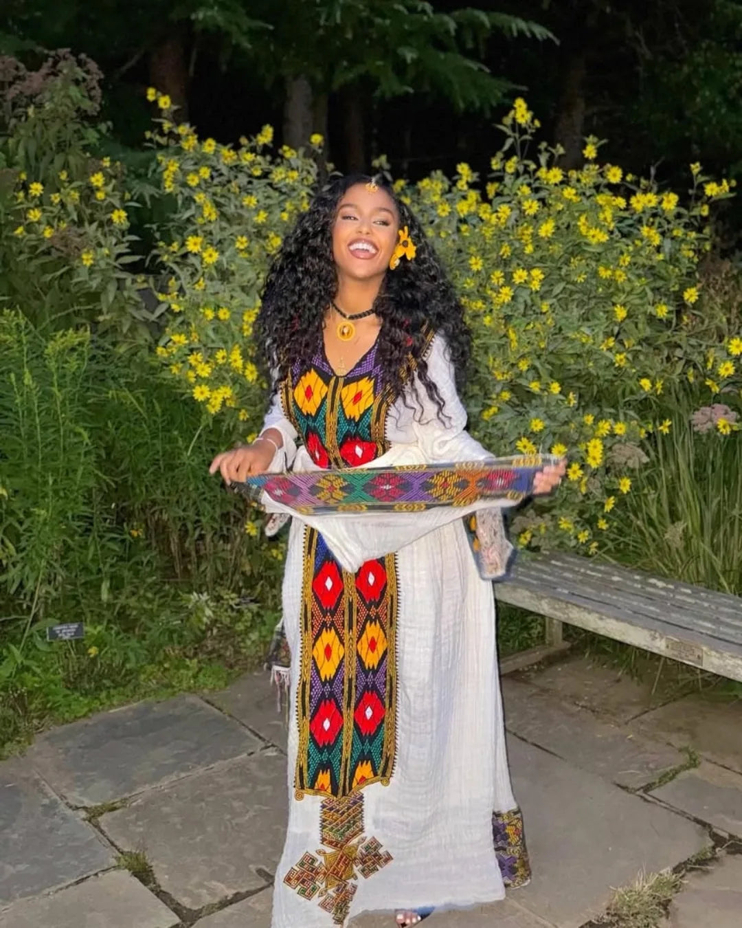 Woman in a long, patterned dress holding a colorful tray outdoors with flowers and a bench in the background. axum habesha kemis ethgebya
