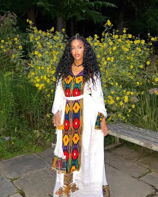 Woman in a colorful traditional outfit standing in a garden with yellow flowers and a bench. axum habesha kemis ethgebya