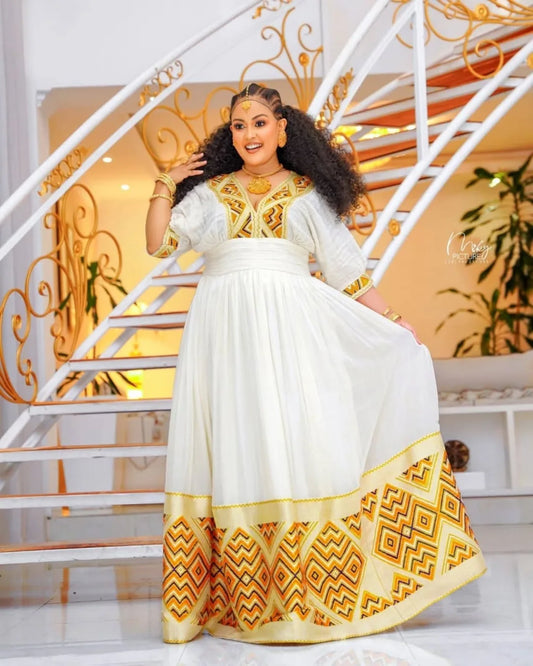 Woman in a white and gold traditional outfit standing on a staircase. Orange & Yellow Zuria Habesha Kemis Modern Ethiopian Traditional Dress Ethgebya