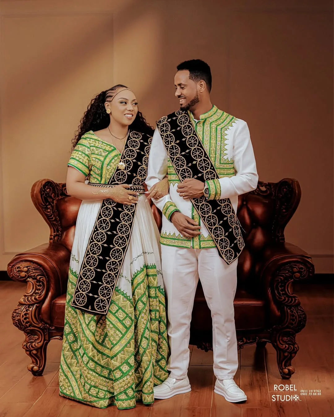 Couple in traditional attire standing in a room with a wooden floor and brown walls.