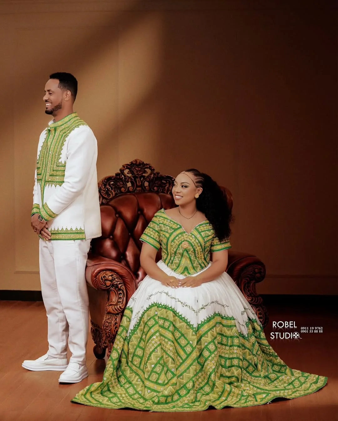 Man and woman in traditional green and white attire posing in a room with a wooden chair. ethiopian and eritrean ethgebya