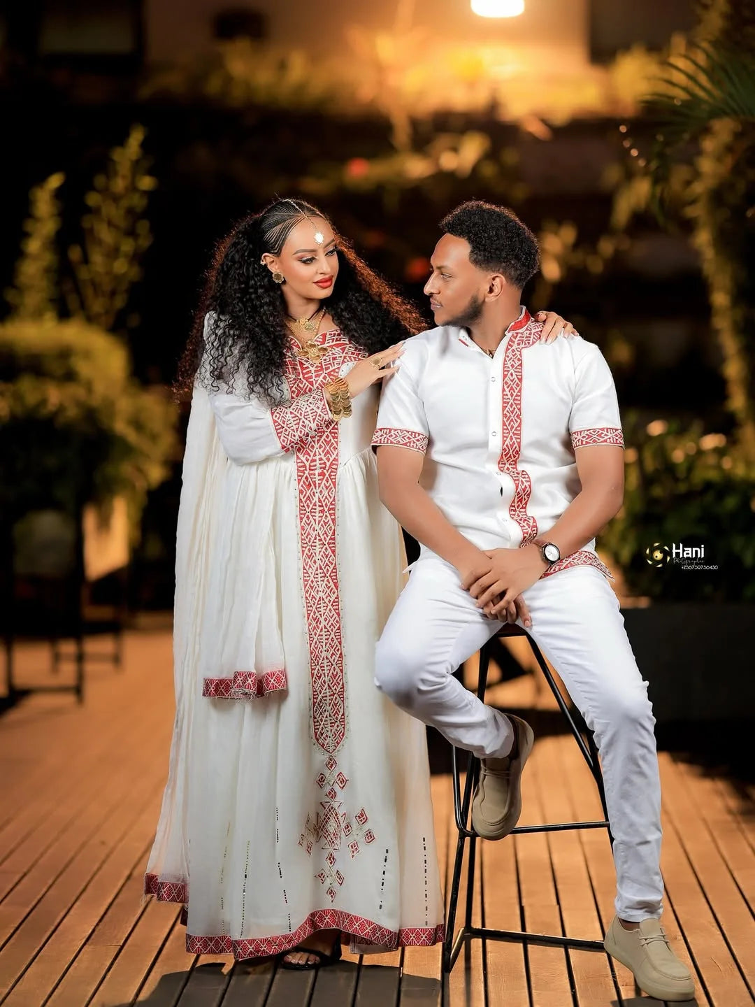 Couple in traditional attire sitting together on a wooden floor with plants in the background Red Ethiopian Couple Set ethgebya