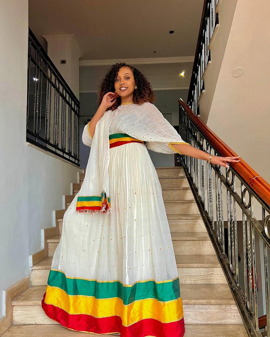 Woman in a white dress with colorful hem standing in a home interior. Green, Yellow & Red Habesha Zuria Kemis Modern Ethiopian Dress ethgebya gebeya