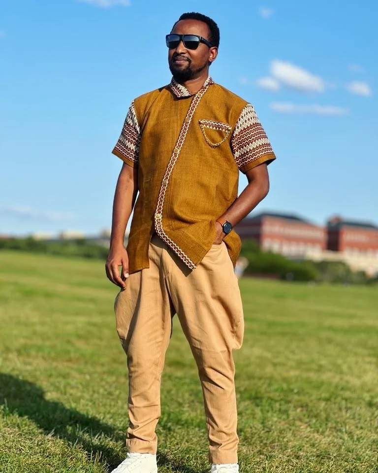 Man wearing a mustard-colored traditional outfit with sunglasses, standing on grass with a building in the background. rown Modern Habesha Men’s Shirt Stylish Ethiopian Traditional Wear ethgebya gebeya usa habesha men