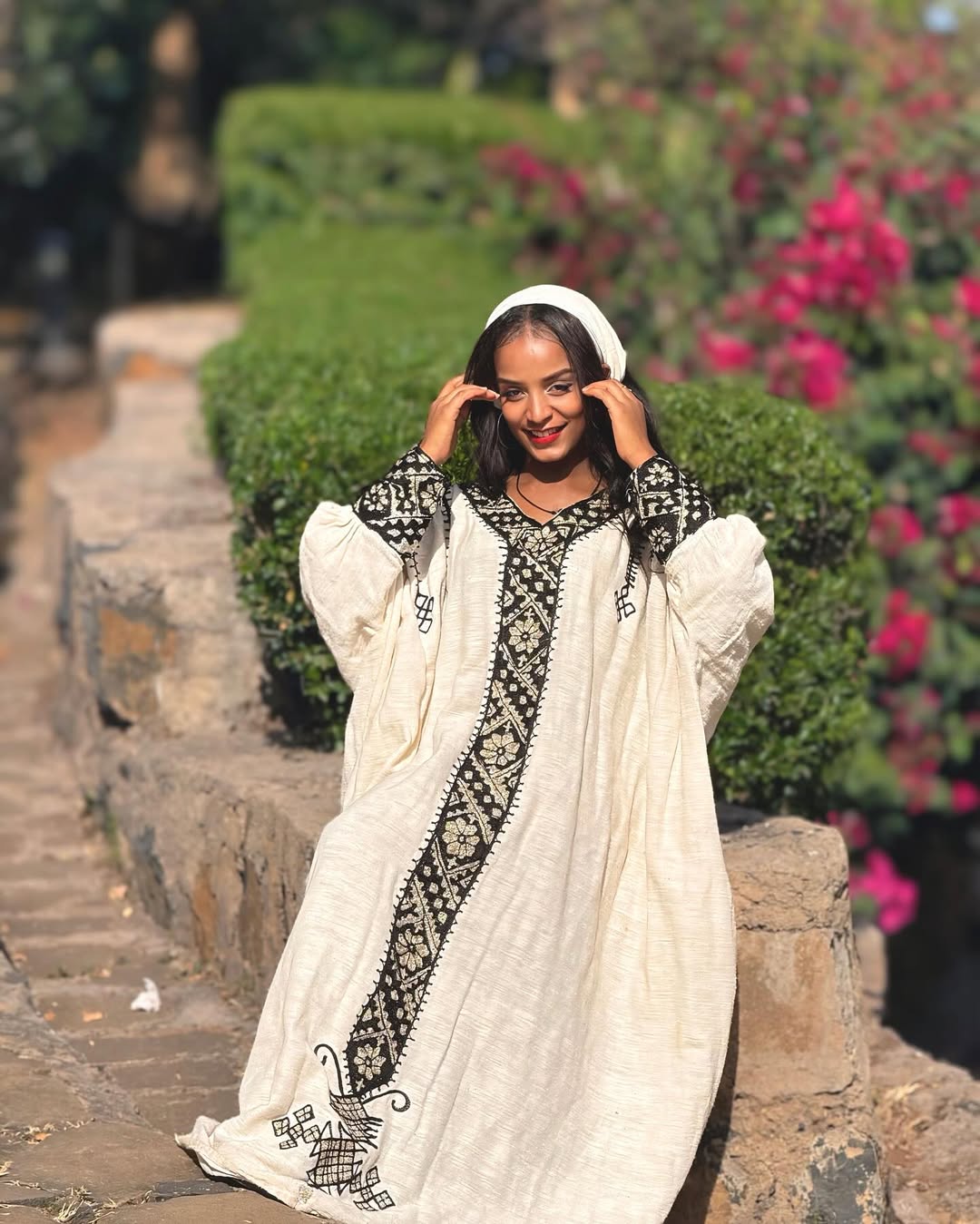 Woman in a white and black embroidered traditional outfit standing outdoors with greenery and flowers in the background. Gonder Habesha Kemis New Relaxed Ethiopian Traditional Dress ethgebya gebeya usa gondar habesha kemis