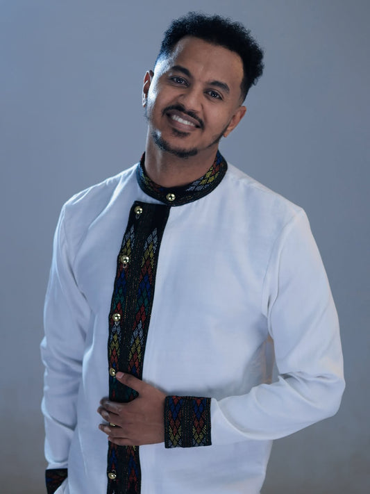 Man wearing a traditional outfit with a white shirt and colorful embroidery on a gray background Ethiopian Men’s Shirt | Handwoven Tilf Embroidery New Habesha Fashion