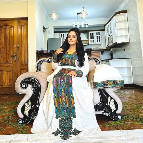Woman in a colorful traditional outfit sitting on an ornate chair in a home setting.