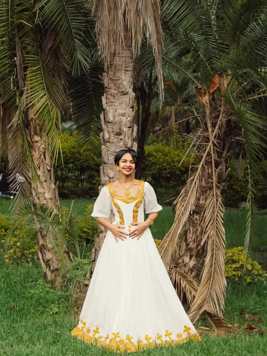 Woman in a white and yellow dress standing in a garden with palm trees. Gold Zuria Habesha Kemis Modern Ethiopian Traditional Dress ethgebya gebeya usa habesha kemis