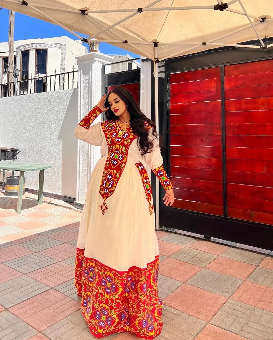Woman in a traditional outfit with red and gold patterns standing outdoors. Stunning Red Zuria Habesha Kemis Handcrafted Ethiopian Dress  Ethiopian artisanal dress, Habesha style, Habesha fashion, Ethiopian cultural wear, Eritrean cultural attire, Habesha modern outfit, Ethiopian classic dress, Ethiopian embroidery fashion, Habesha traditional clothing, Ethiopian traditional outfits for women, Habesha chic dress, Ethiopian ethnic dress, Habesha inspired fashion, Ethiopian custom made dress, modern Kethgbeya