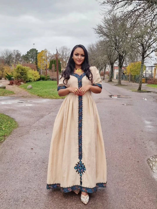 Woman in a long, beige dress with blue embroidery standing on a street. Beige Modern Zuria Habesha Kemis Stylish Ethiopian Traditional Dress ethgebya gebeya habesha kemis