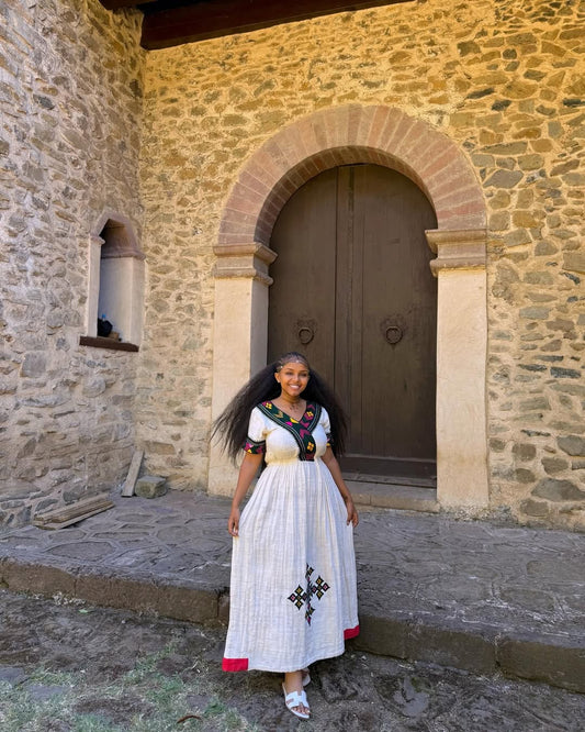 Woman in a white dress with black patterns standing in front of a stone building with an arched door.  Multicolor Gondar Habesha Kemis Modern Ethiopian Traditional Dress ethgebya gebeya