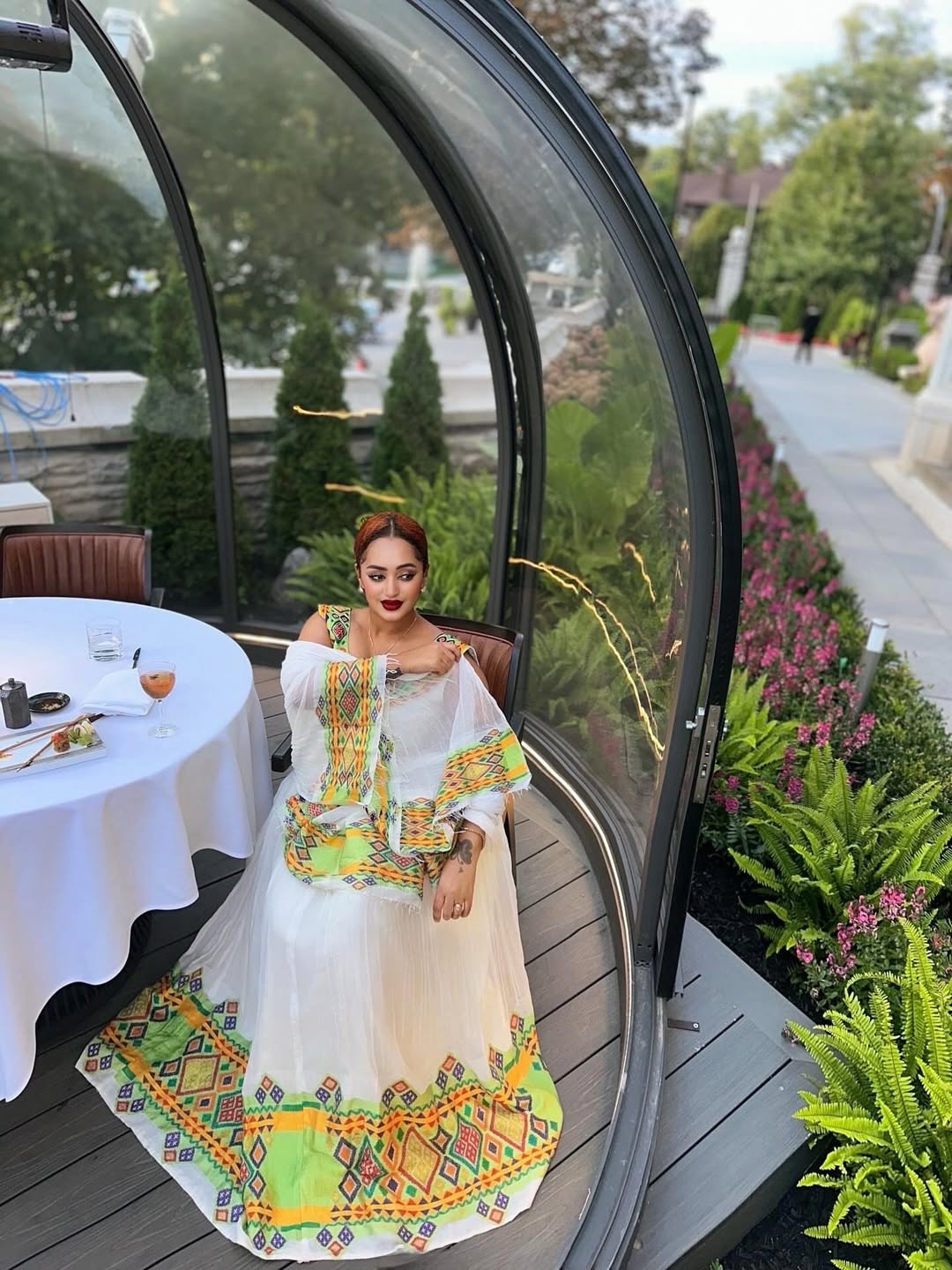 Woman in a colorful dress standing in front of a glass door with a garden view. Beautiful Modern Zuria Kemis | Elegant Ethiopian Traditional Dress