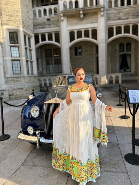 Woman in a white dress with colorful patterns standing in front of a vintage car and a historic building. Beautiful Modern Zuria Kemis | Elegant Ethiopian Traditional Dress
