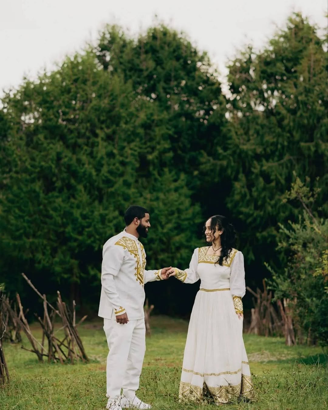 Couple in traditional white outfits with gold details standing in a field with trees in the background. Golden Habesha Couple Outfit Modern Ethiopian Matching Set ethgebya gebeya