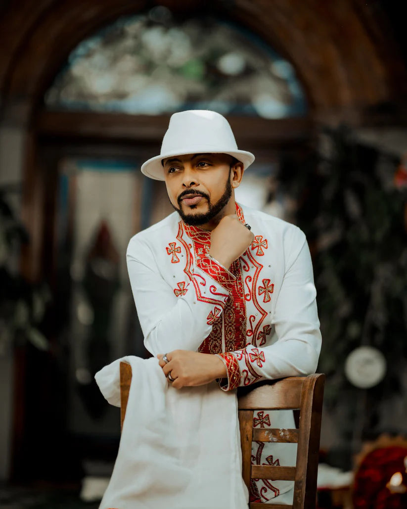 Man wearing a white embroidered outfit with red patterns and a white hat, sitting on a wooden chair. ethiopian and eritrean mens traditional wear ethgebya