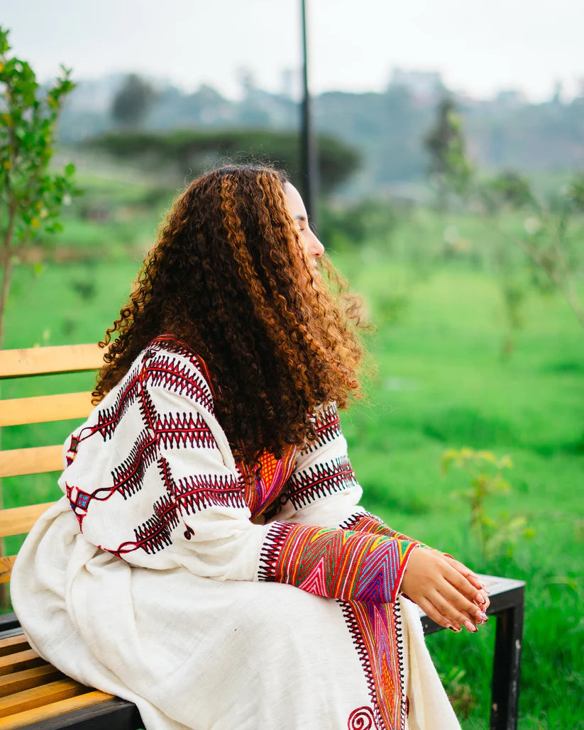 Woman sitting on a bench wearing a colorful shawl with a green landscape in the background gondar habesha kemis ethgebya
