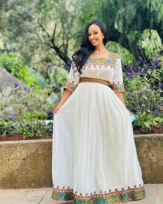 Woman in a white dress with colorful embroidery standing outdoors in a garden.  Light Green & Gold Zuria Habesha Kemis Ethgebya