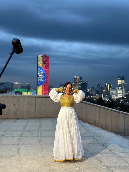 Woman in a yellow top and white skirt on a rooftop with city skyline in the background Gold Zuria Habesha Kemis New Elegant Modern Ethiopian Dress ethgebya gebeya habesha kemis