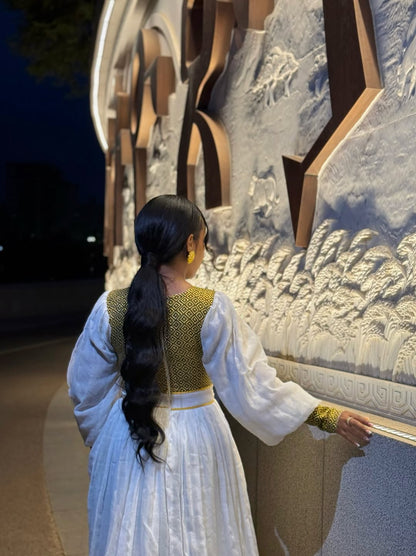 Woman in a white dress with a green top standing in front of a decorative wall. Gold Zuria Habesha Kemis New Elegant Modern Ethiopian Dress ethgebya gebeya habesha kemis