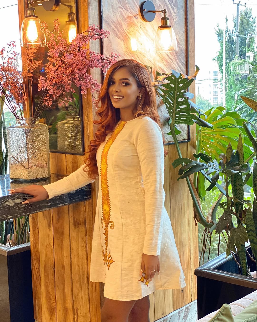 Woman in a white traditional outfit with gold details standing  Modern Yellow Lightweight Short Habesha Kemis Ethiopian Dress ethgebya habesha kemis gebeyaindoors with plants and decor.