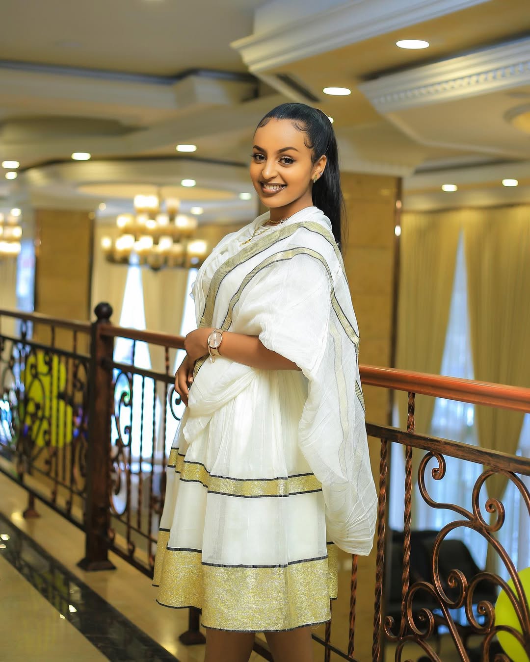Woman in a white and gold traditional outfit standing in an elegant interior setting. Simple Short Golden Habesha Dress Modern Summer Ethiopian Attire ethgebya gebeya usa habesha kemis
