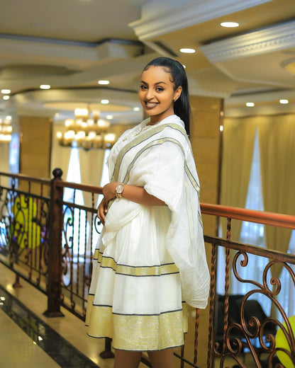 Woman in a white and gold traditional outfit standing in an elegant interior setting. Simple Short Golden Habesha Dress Modern Summer Ethiopian Attire ethgebya gebeya usa habesha kemis