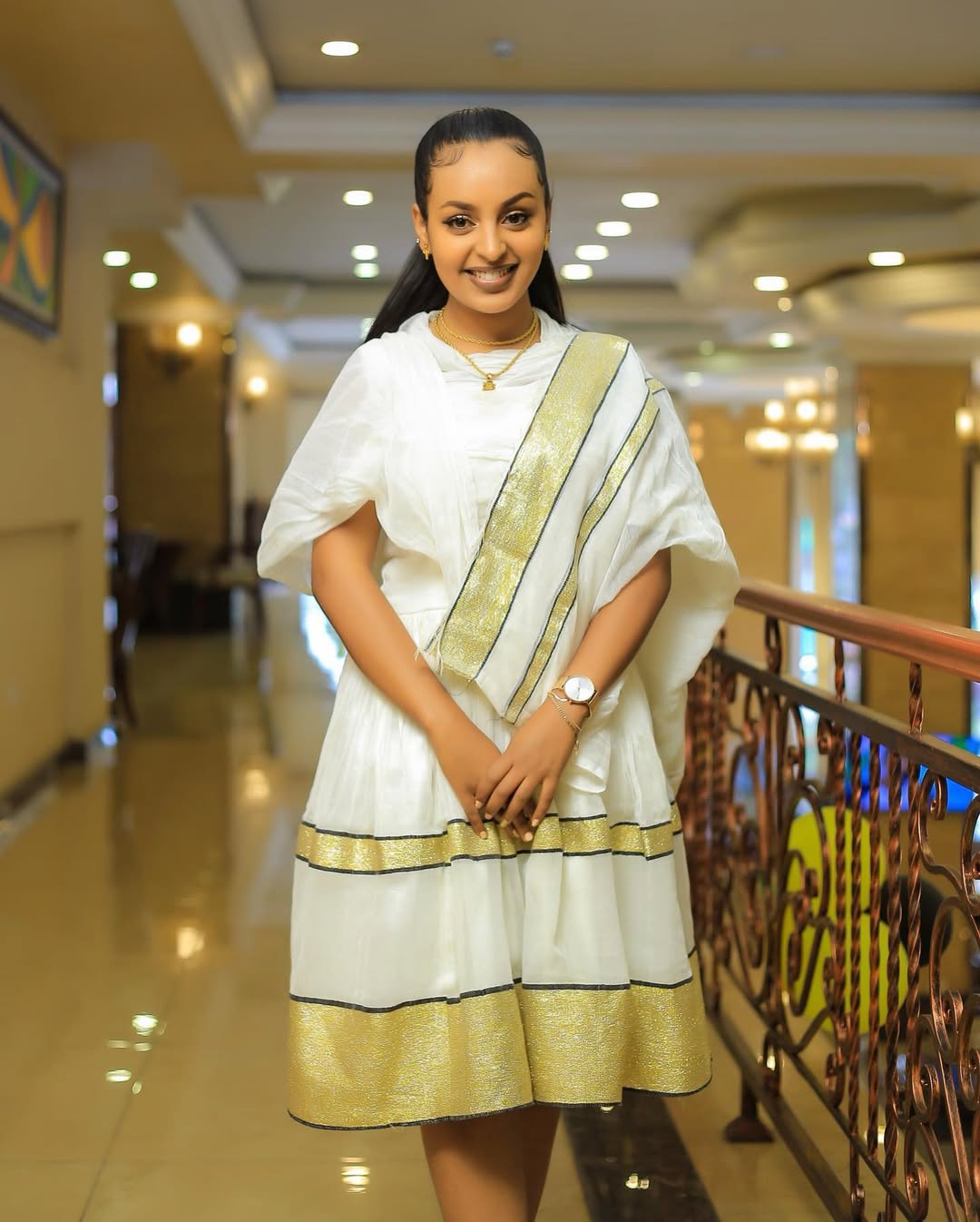 Woman in a white and gold traditional outfit standing in a well-lit indoor setting. Simple Short Golden Habesha Dress Modern Summer Ethiopian Attire ethgebya gebeya usa habesha kemis