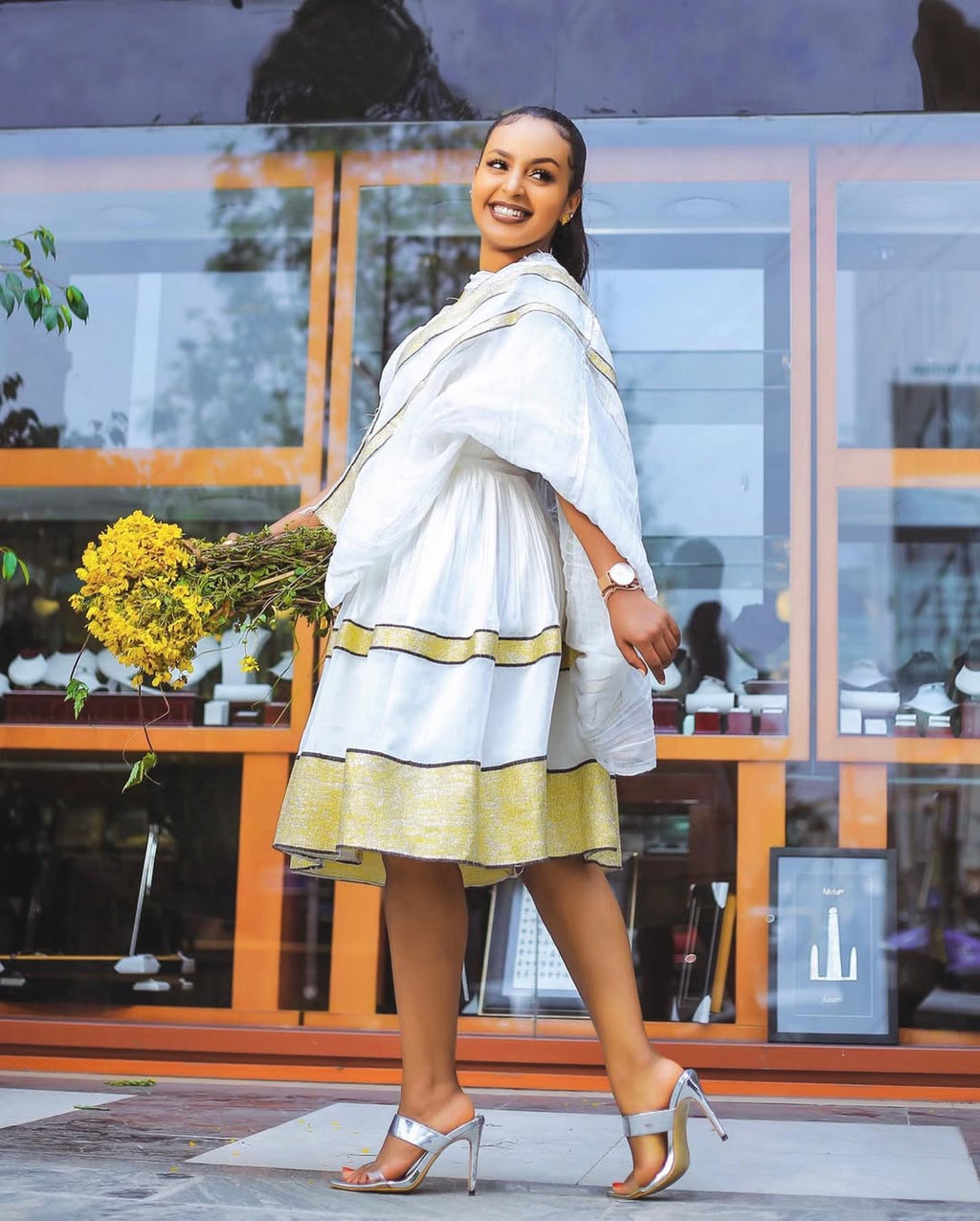 Woman in a white and yellow dress holding flowers in front of a store window. Simple Short Golden Habesha Dress Modern Summer Ethiopian Attire ethgebya gebeya usa habesha kemis
