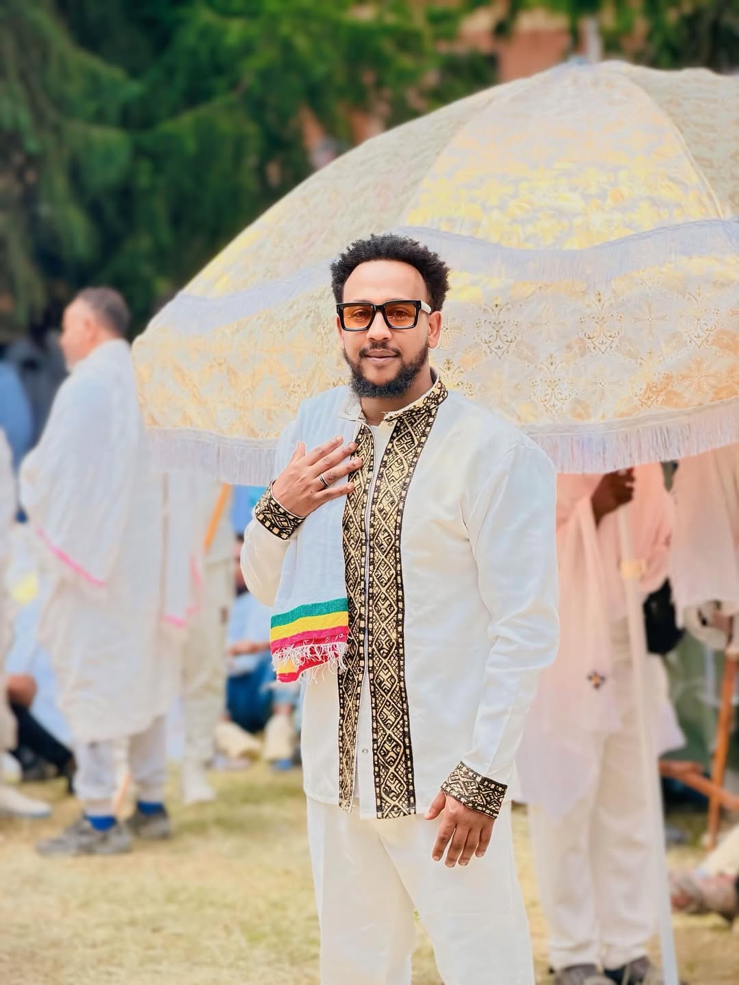 Man in traditional outfit with colorful accents standing under a decorative umbrella outdoors. Gold Habesha Men’s Shirt Modern Ethiopian Traditional Wear ethgebya gebeya
