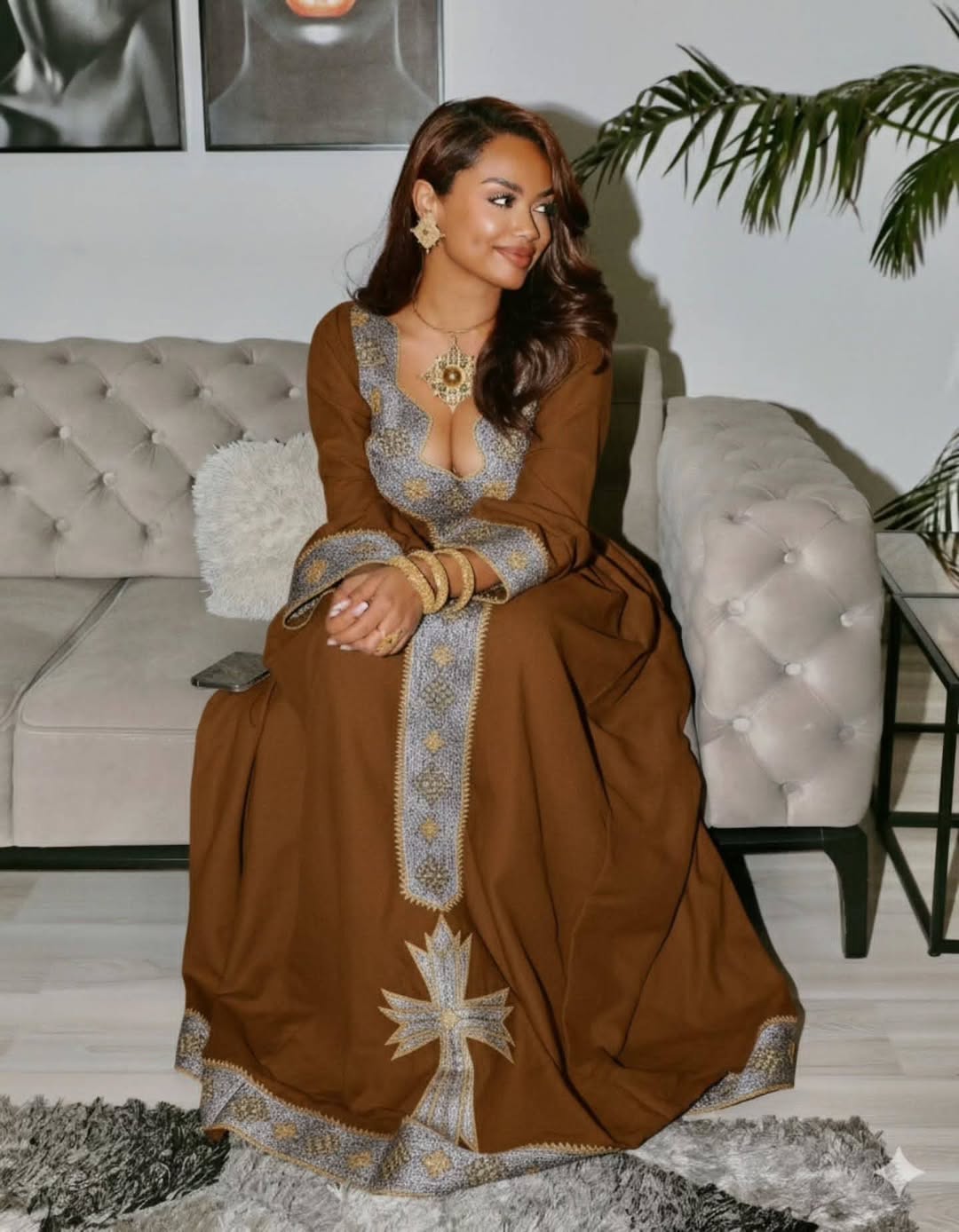 Woman in a brown dress with intricate patterns sitting on a couch in a living room. Brown Dominant Zuria Habesha Kemis Modern Ethiopian & Eritrean Dress Ethiopia Gebeya Ethgebya