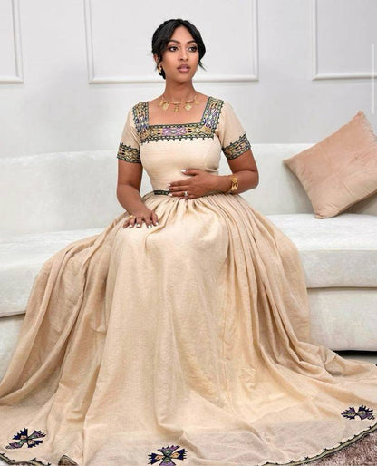 Woman in a beige saree with intricate designs sitting on a white couch. New Beige Zuria Habesha Kemis Modern Handcrafted Ethiopian Dress ethgebya gebeya