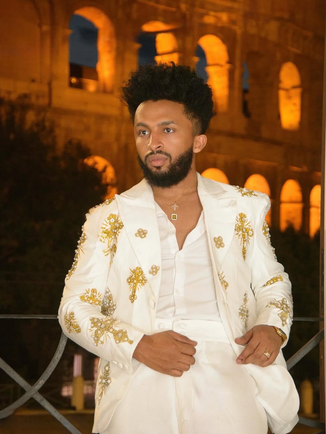 Man in a white embroidered suit standing in front of an illuminated building. Gold Tilf Habesha Men’s Shirt Modern Ethiopian Traditional Wear ethgebya gebeya