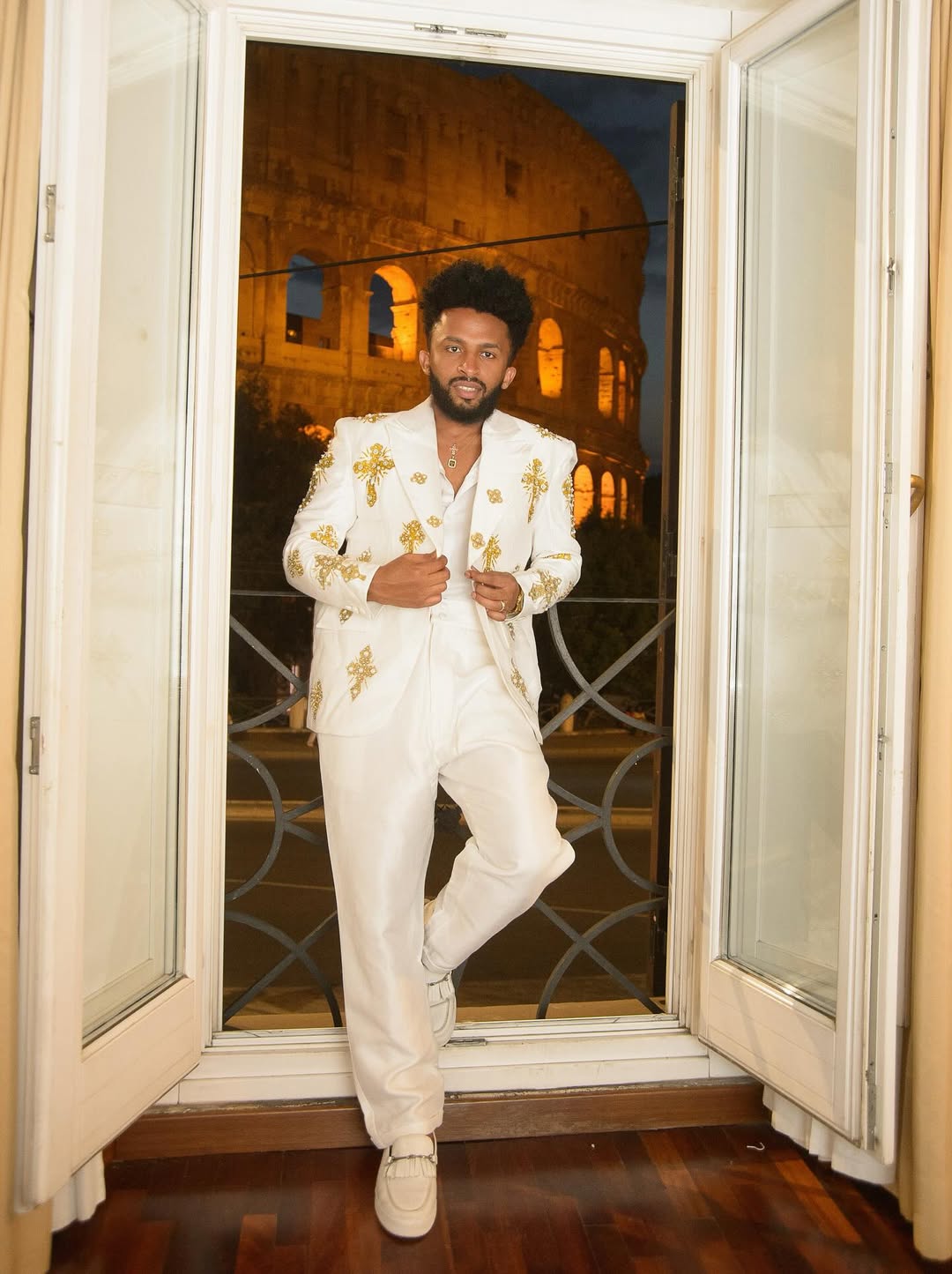 Man in a white suit with gold accents standing in front of a window with a view of the Colosseum. Gold Tilf Habesha Men’s Shirt Modern Ethiopian Traditional Wear ethgebya gebeya