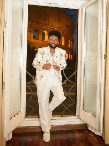 Man in a white suit with gold accents standing in front of a window with a view of the Colosseum. Gold Tilf Habesha Men’s Shirt Modern Ethiopian Traditional Wear ethgebya gebeya