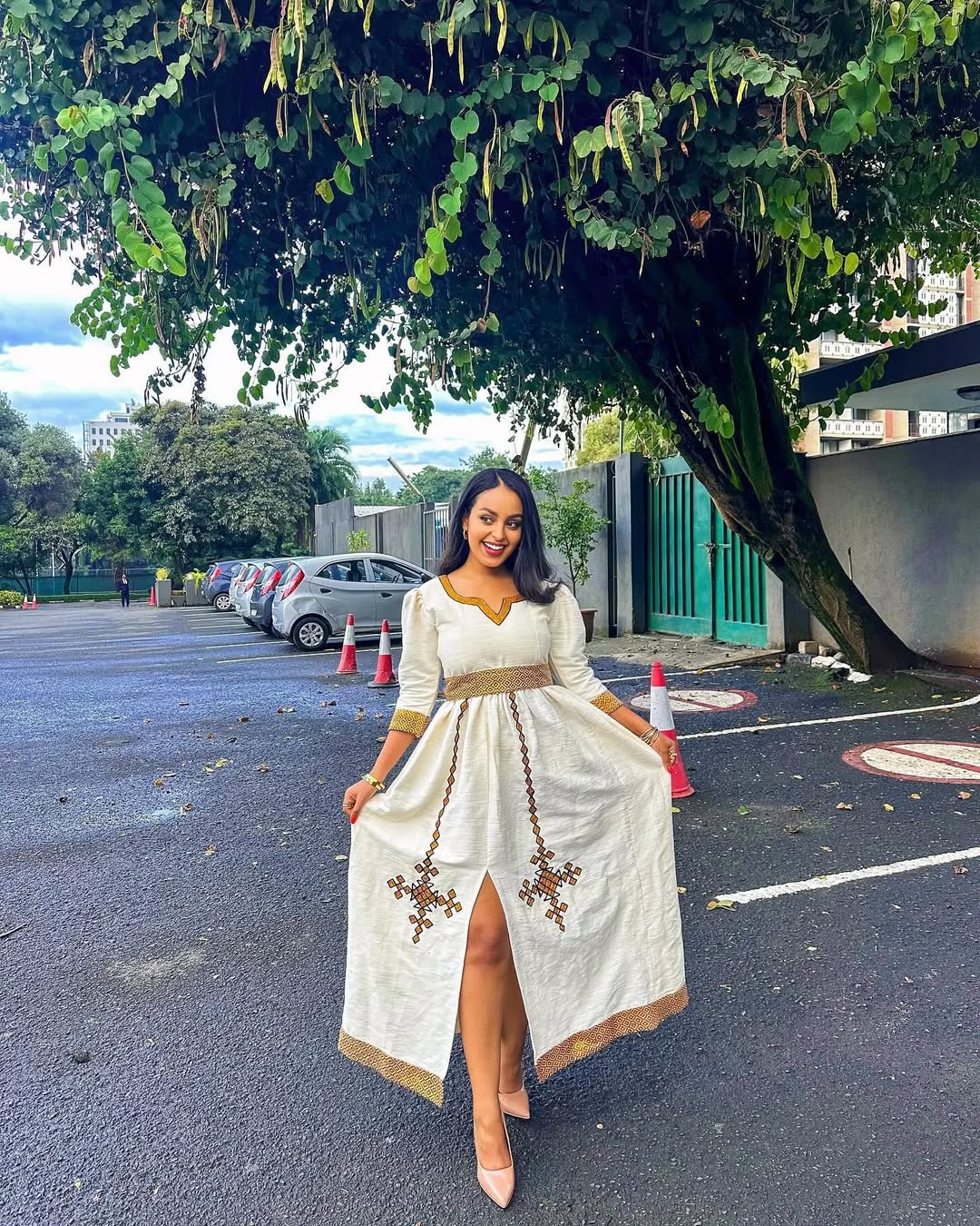 Woman in a white dress with gold accents standing on a street with trees and buildings in the background. Simple Golden Zuria Habesha Kemis New Elegant Ethiopian Dress ethgebya gebeya